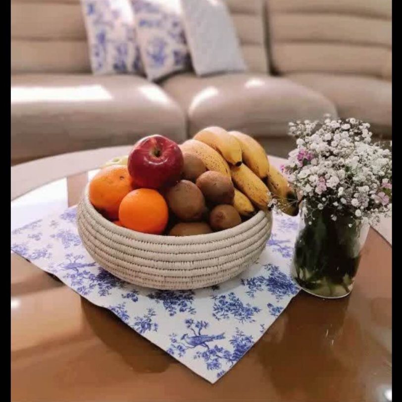 ظرف میوه حصیری روی میز قرار دارد درون آن میوه پرتقال و موز و انار و کیوی قرار دارد